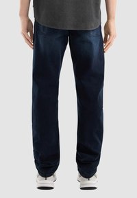 Vaqueros de mezclilla azul oscuro con un corte relajado, que cuentan con bolsillos traseros y un tobillo ligeramente ajustado. Llevados con zapatillas blancas.