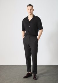 Chemise noire à rayures côtelées à manches courtes avec boutons, associée à un pantalon noir ajusté et des chaussures habillées bordeaux foncé, sur fond neutre.