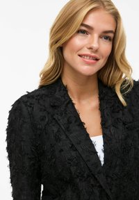 Black blazer with textured fabric featuring frayed detailing, single button closure, and a notched collar, worn over a white top.