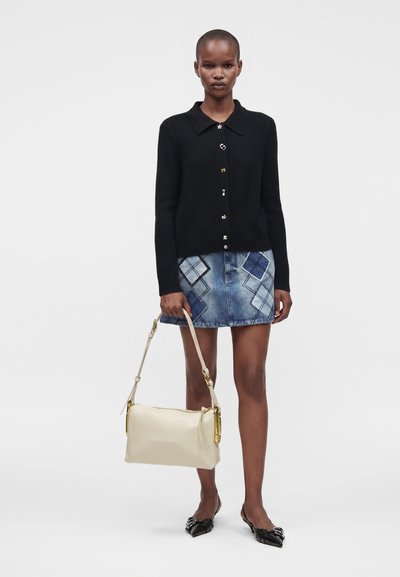 Femme portant un cardigan noir boutonné, une jupe en jean bleue à motifs, des ballerines noires pointues, et tenant un sac à main beige par la anse.