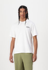 Fehér pamut póló rövid ujjakkal, kerek nyakkal és mellkasi logóval, amely a "DICKIES" feliratot viseli. Olívazöld nadrággal viselik, amelyen látható zsebek találhatók.
