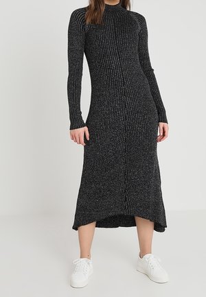 Kobieta w długiej, prążkowanej, ciemnoszarej dzianinowej sukience z długimi rękawami i białych tenisówkach, stojąca na tle jednolitego jasnego tła.