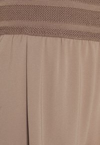 Beige jurk met een gestructureerde elastische tailleband, en met plooien in een gladde stof, die een gestructureerd maar vloeiend silhouet creëren.