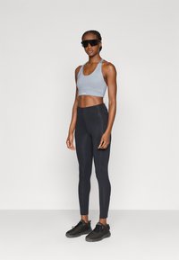 Αθλητικό σουτιέν ανοιχτό γκρι με λαιμόκοψη scoop, μαύροι ψηλόμεσοι κολάν και μαύρα αθλητικά παπούτσια. Μοντέλο που φοράει σκούρα γυαλιά ηλίου.