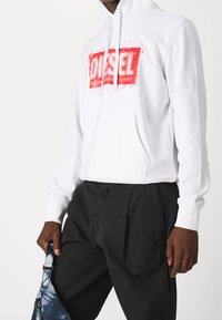 Vit hooded sweatshirt med framficka, med ett rött logotryck. Lager ovanpå mörka byxor, hålls av en hand med en blå väska.