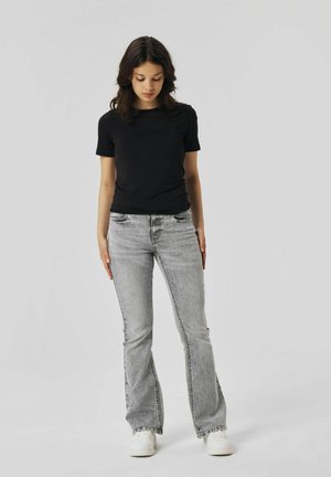 T-shirt nera aderente abbinata a jeans svasati grigi. I jeans presentano una texture scolorita; il modello indossa sneakers bianche, in piedi con le mani ai fianchi.