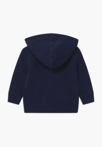 Felpa blu navy con texture morbida, dotata di cappuccio con cordino e polsini a coste. Dettagli delle cuciture lungo la schiena per dare struttura.