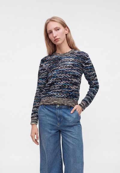 Femme aux longs cheveux blonds portant un pull en tricot multicolore et un jean bleu à jambes larges, debout avec une main dans la poche, sur un fond uni.