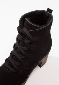 Bottine en daim noir avec talon épais, fermeture à lacets à l'avant et doublure intérieure moelleuse, vue en angle de dessus sur fond blanc.