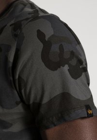 Tričko s kamuflážovým vzorem v tmavě šedé a černé, vyrobené z měkkého materiálu. Disponuje designem s krátkým rukávem, šitým lemem a štítkem s logem.
