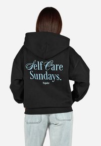 Čierna mikina s kapucňou, ktorá na chrbte obsahuje veľký svetlomodrý text s nápisom "Self Care Sundays." a menším písmom "Pegador."