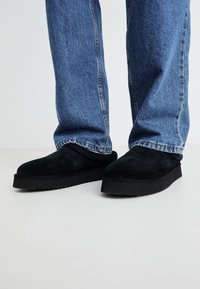 Zapatos negros de ante de tipo slip-on con suela de goma plana, combinados con jeans de mezclilla azul de pierna recta. Diseño minimalista con una textura suave.