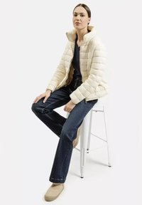 Jasnobeżowa puchowa kurtka z wysokim kołnierzem, noszona na ciemnej koszuli, w komplecie z ciemnymi dżinsami i beżowymi butami, siedząca na białym stołku.