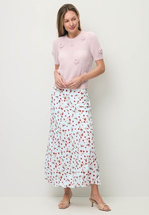 Kobieta w różowym sweterku z krótkim rękawem ozdobionym kwiatowymi detalami oraz białej długiej spódnicy w czerwone truskawki, beżowe sandały na obcasie.