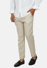 Pantaloni di lino beige con una vestibilità casual, dotati di tasche laterali e orli arrotolati, abbinati a scarpe nere e una camicia bianca con bottoni.