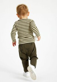 Gestreept overhemd met lange mouwen in olijf en crème, gecombineerd met olijfgroene broek met een bladenprint en witte sneakers. Kind dat wegkijkt.