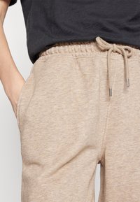 Beżowe spodnie dresowe o miękkiej fakturze, z elastycznym pasem, sznurkiem ze stalowymi końcówkami oraz kieszeniami po bokach. Zestawione z ciemnym T-shirtem.