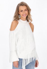 Witte gebreide trui met off-shoulder ontwerp, franjes langs diagonale patronen, geribde hoge hals en belmouwen. Gepaard met lichtblauwe jeans.