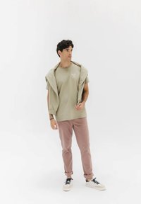 T-shirt di cotone verde chiaro con maniche corte e logo bianco, abbinata a pantaloni chino rosa e sneakers bianche, con una felpa grigia appoggiata sulle spalle.