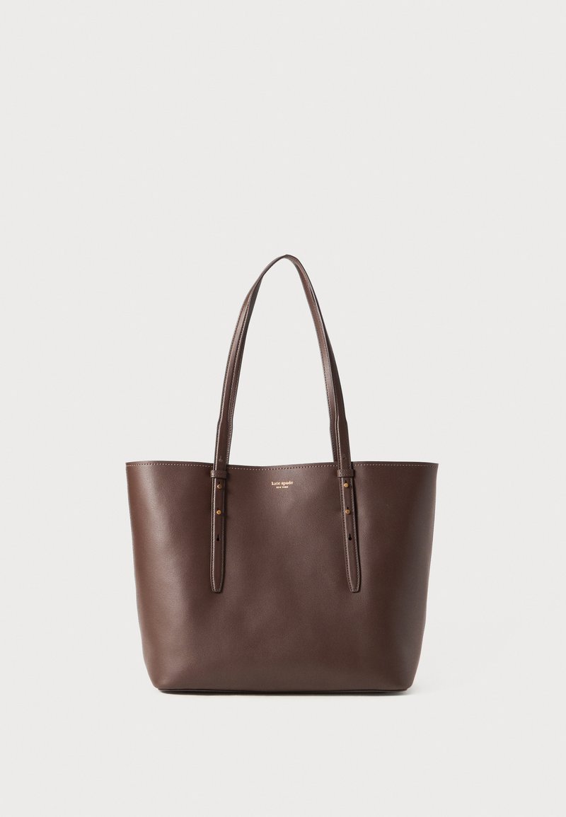 Brown leather tote bag with two long handles, featuring a smooth texture, structured shape, and subtle stitch detailing. Gold logo on front.