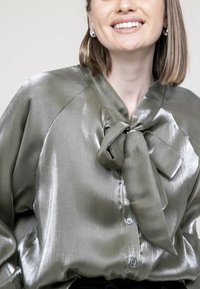 Blouse en satin vert olive avec un détail de nœud, fermeture à boutons et une texture brillante et lisse. Présente une coupe décontractée et un col.