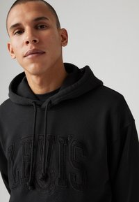 Black hoodie made of cotton blend, featuring a large embossed "LEVI'S" logo and drawstring hood, with a soft texture and relaxed fit.