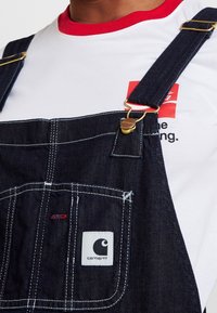 Pantalones de mezclilla oscuros con herrajes dorados, costuras blancas y un parche de logo de Carhartt; llevados sobre una camisa blanca con un collar rojo y detalles del logo.