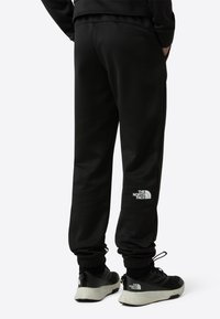 Čierne teplákové nohavice s elastickými manžetami a bočnými vreckami, s bielym logom na ľavej strane. Nosené s čiernymi a sivými športovými topánkami.