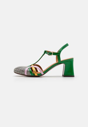 Sandale à talon vert avec bout géométrique noir et blanc, brides dorées et roses, et boucle à la cheville sur fond blanc.