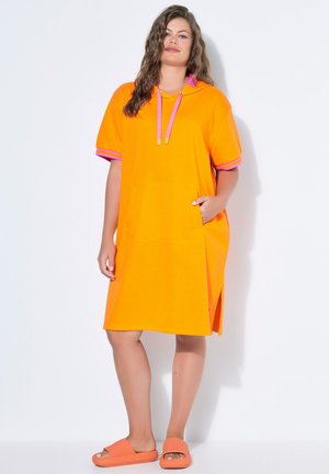 Frau steht und trägt ein orangefarbenes, knielanges Hoodie-Kleid mit pinken Akzenten sowie passende orangefarbene Slide-Sandalen vor weißem Hintergrund.