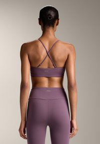 Vijolična športna modrček z križnimi naramnicami in logotipom na hrbtni strani, v kombinaciji z vijoličnimi hlačami z visokim pasom. Gladka, atletska tekstura tkanine.