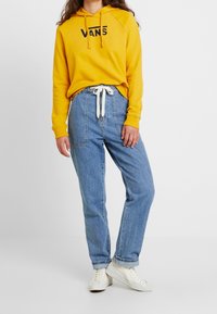 Felpa gialla con logo "VANS" nero, abbinata a jeans di denim blu chiaro. Il look è completato da un laccio bianco e sneakers bianche casual.