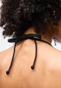 Personne à cheveux bouclés et à la peau foncée, portant une bride de soutien-gorge noire attachée en nœud derrière le cou, avec une boucle d'oreille en or.