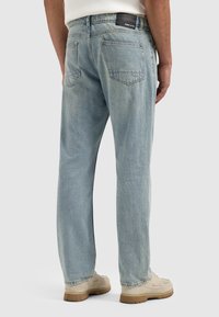 Man in lichtblauwe rechte spijkerbroek en beige schoenen, staande met de handen ontspannen langs de zij tegen een effen achtergrond.