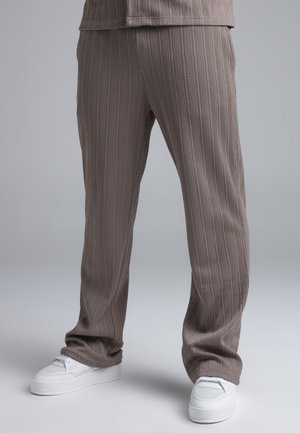 Személy laza, bordázott, törtfehér színű nadrágot visel, fehér tornacipővel, és egy egyszínű világosszürke háttér előtt áll.