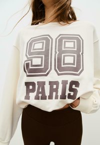 Felpa crema con grande stampa grigio scuro "98" e "PARIS". Presenta maniche lunghe, polsini ripiegati e una vestibilità rilassata. Abbinata a leggings marroni.