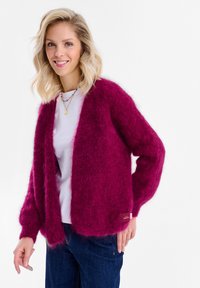 Uśmiechnięta kobieta o falowanych blond włosach, ubrana w puszysty karminowy kardigan, biały t-shirt, warstwowe naszyjniki i ciemnoniebieskie jeansy na jednolitym tle.