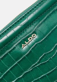 Cuir vert à texture crocodile avec logo ALDO doré et couture de fermeture éclair visible le long du bord d'un sac à main ou d'un portefeuille.