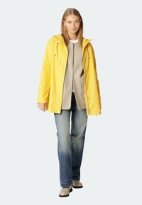 Geel waterdichte jas met capuchon, ritszakken en drukknoopsluiting, gelaagd over een beige shirt, gecombineerd met blauwe flared jeans en beige laarzen.