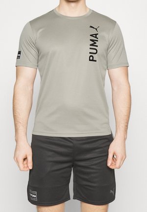 Mężczyzna w jasnoszarej koszulce z krótkim rękawem Puma z pionowym logo i czarnych szortach Puma z naszywkami z logo.