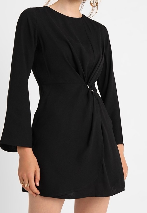 Modèle portant une robe noire à manches longues, avec un design avant torsadé et des accents argentés à la taille, se tenant contre un fond clair.