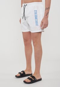 Witte zwembroek met een trekkoord in de taille, met de woorden "Bikkembergs" in blauw en lichtoranje tekst langs de zijkant. Gekozen met zwarte slipper sandalen.