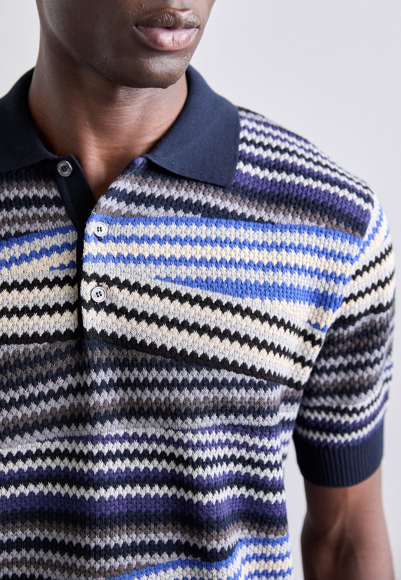 Polo a maniche corte con colletto blu navy. Presenta strisce zigzag in blu, crema e nero. Materiale morbido con un pattern a maglia testurizzata.