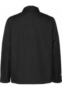 Veste noire à manches longues vue de dos avec un col simple et un tissu lisse, présentant un petit logo réfléchissant près du poignet droit.