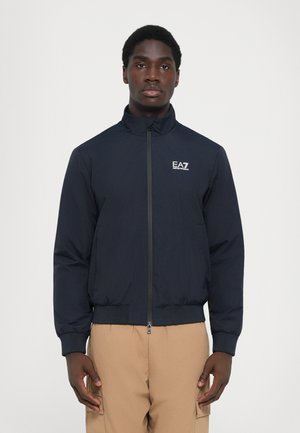 Marine bomberjakke med glidelås, stående krage, elastiske mansjetter og hem. Har en logo på brystet. Båret over beige cargo bukser.