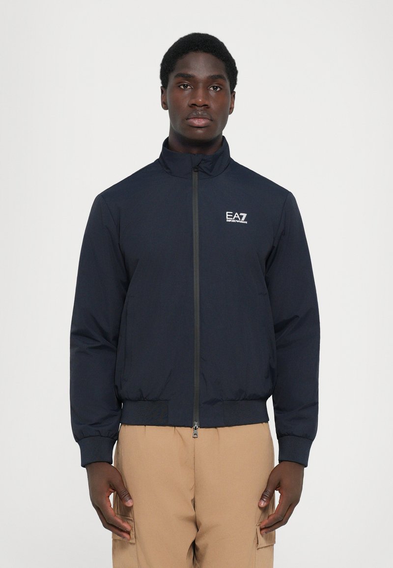 Navy bomber jacket with zip closure, stand-up collar, elastic cuffs, and hem. Features a logo on the chest. Worn over tan cargo pants.