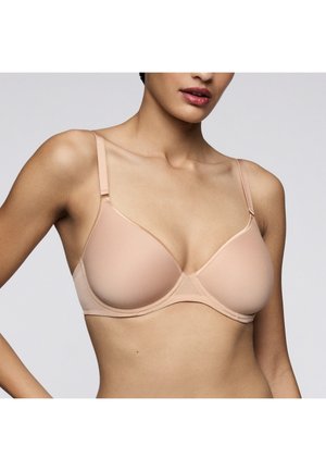 Vrouw draagt een nude kleurige, gladde beugelbeha met verstelbare bandjes tegen een effen lichtgrijze achtergrond.