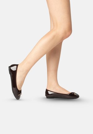 Piernas con bailarinas marrón oscuro brillantes con pequeños lazos, un pie apoyado completamente y el otro ligeramente levantado sobre un fondo liso.