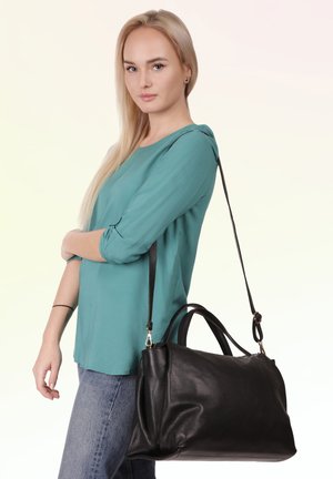 Vrouw met lang blond haar draagt een teal top en spijkerbroek, met een grote zwarte leren schoudertas.