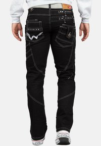 Jeans in denim nero con cuciture bianche, numerose tasche tra cui tasche posteriori decorative, e una cintura piatta con un'etichetta.